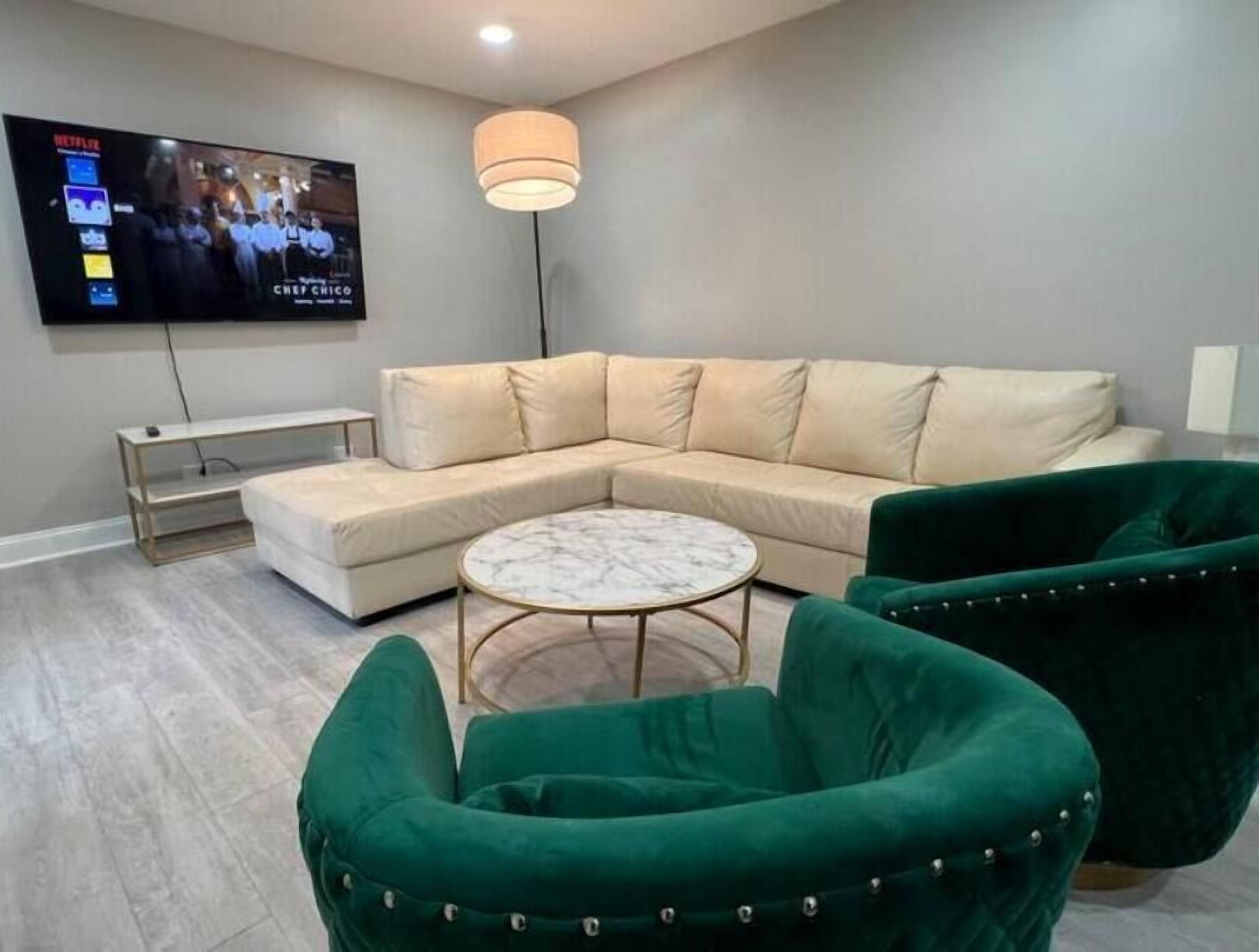 A modern living room with a sectional sofa, a round coffee table, and green armchairs. A flat-screen TV is mounted on the wall.