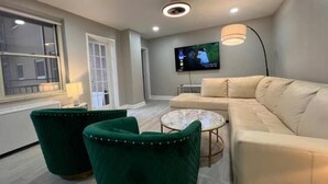 Un salon moderne avec un canapé beige, deux fauteuils verts, une petite table basse ronde, un téléviseur fixé au mur et une fenêtre avec des stores.