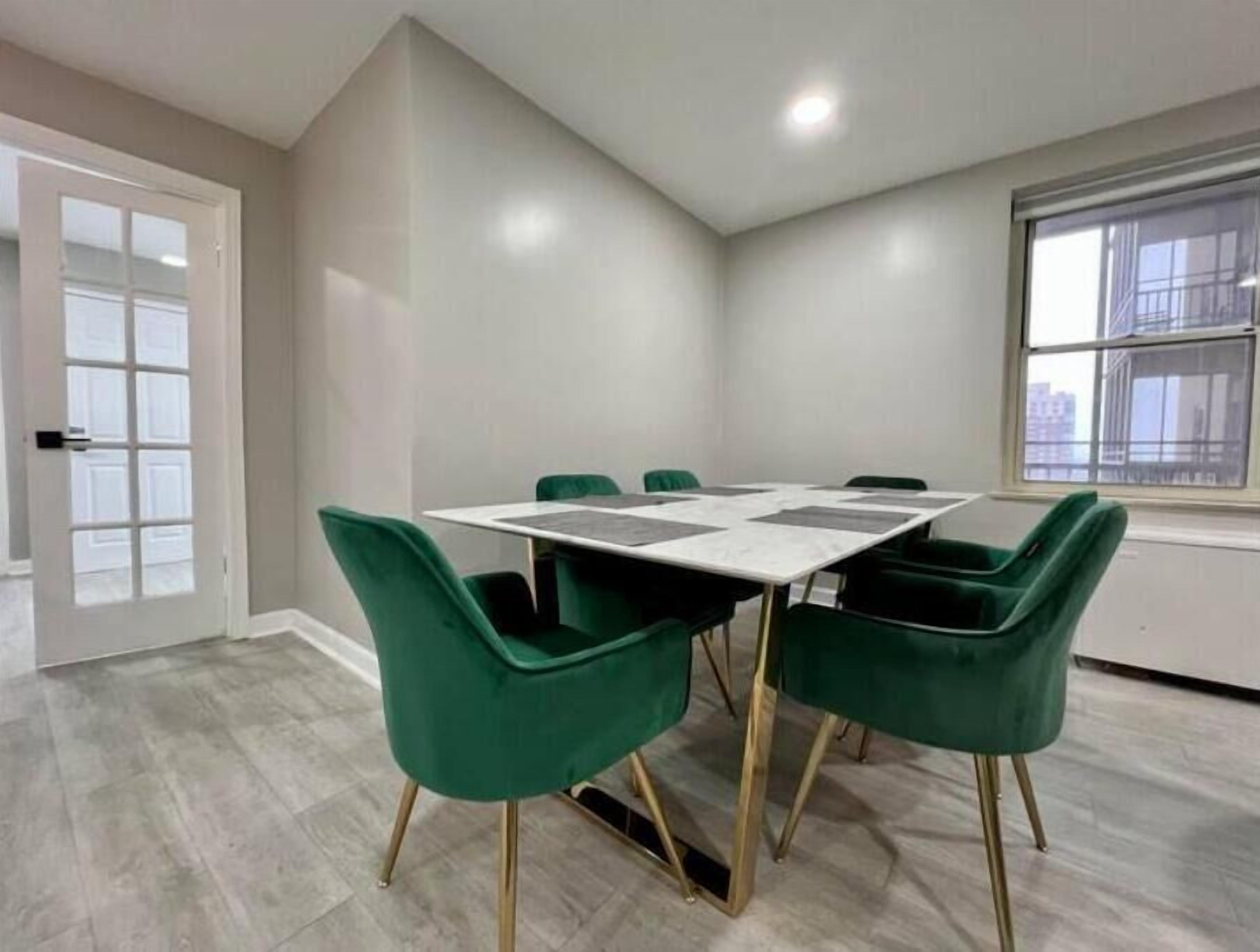 Uma sala de conferências com uma mesa retangular e quatro cadeiras verdes com pés dourados. O espaço possui uma porta branca, uma janela com vista para edifícios e um piso de cor clara.