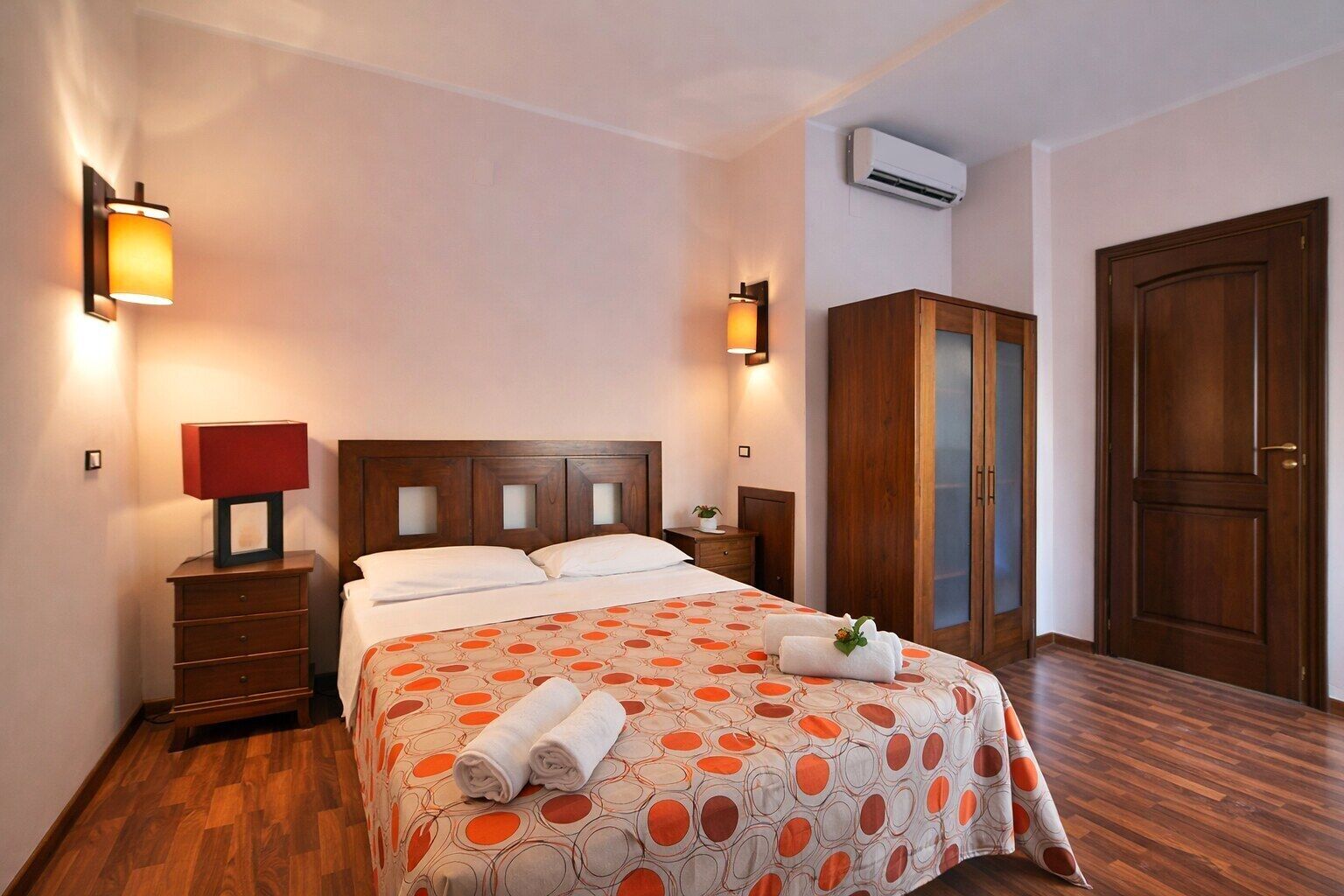 A hotel room with a wooden bed, two bedside tables, a wardrobe, and an air conditioning unit.