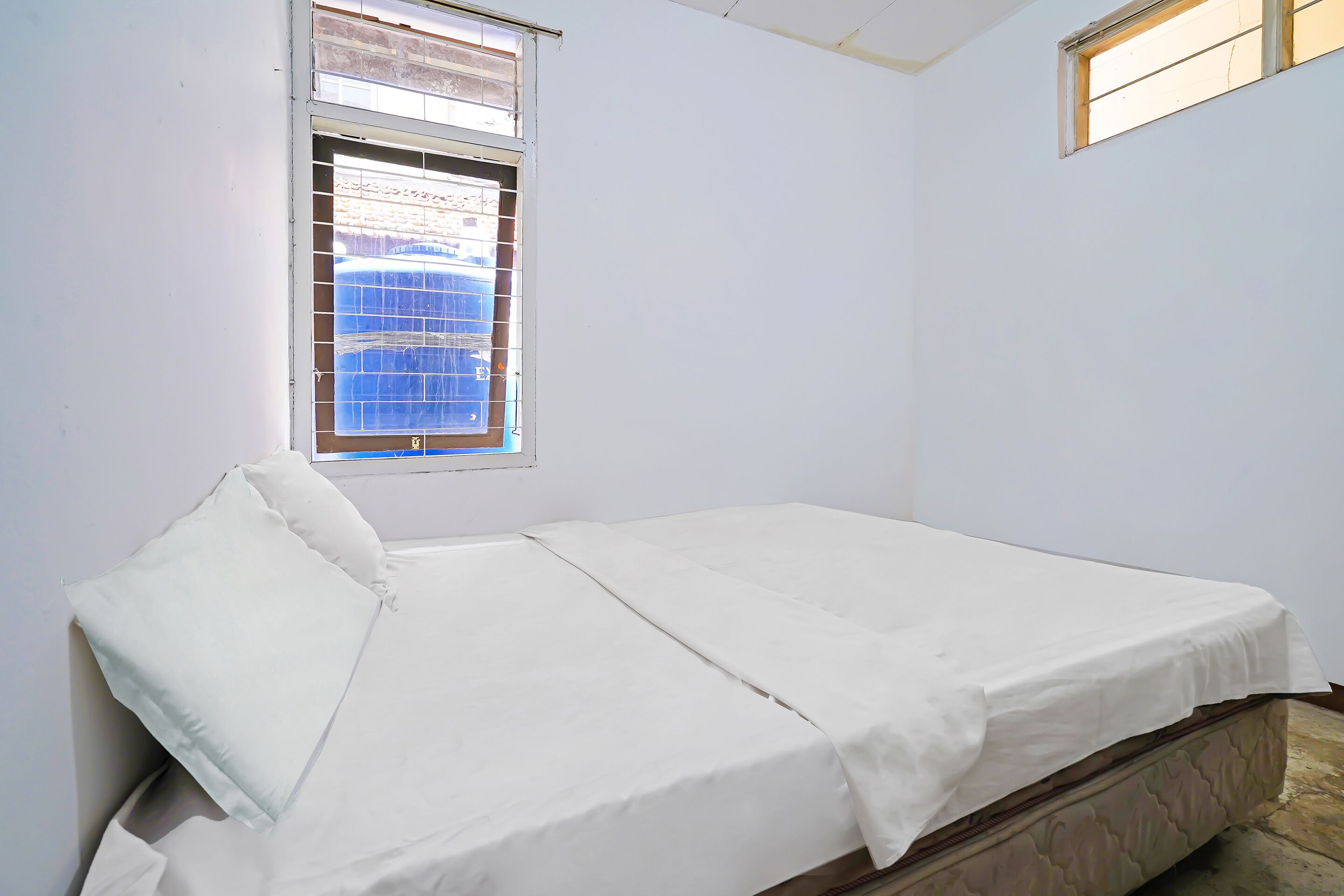 Een enkel bed met een witte matras en kussens, een raam met metalen tralies en een effen witte muur.