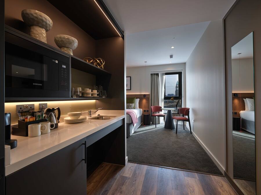 A modern hotel room with a kitchen area, a dining space, and a view of a cityscape.