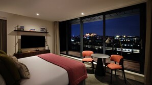 Habitació d'hotel amb un llit, una televisió, una taula amb cadires i una finestra gran que ofereix vistes de la ciutat de nit.