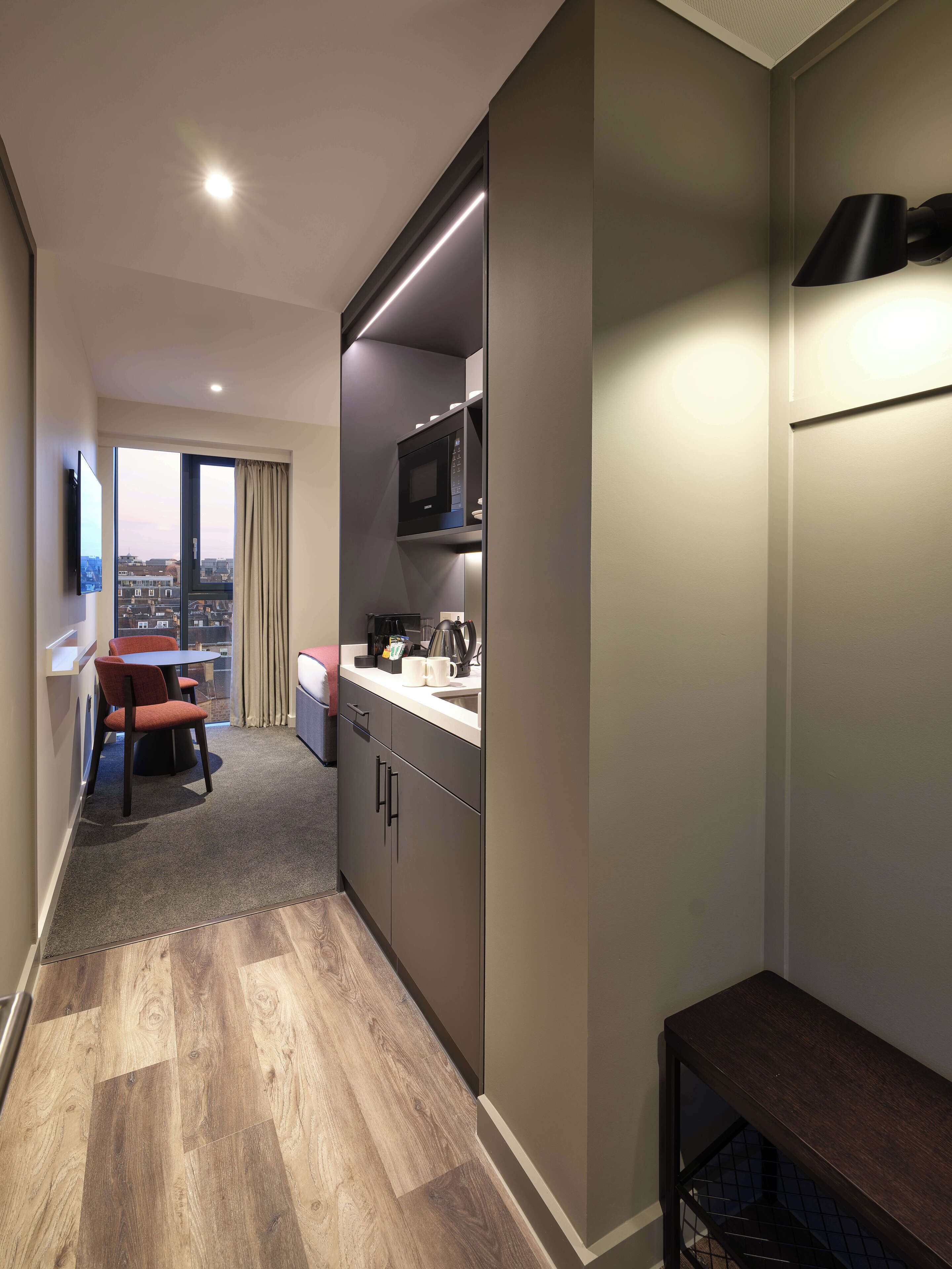 Una habitación de hotel moderna con una zona de cocina, una mesa de comedor con sillas y vistas a la ciudad a través del ventanal.