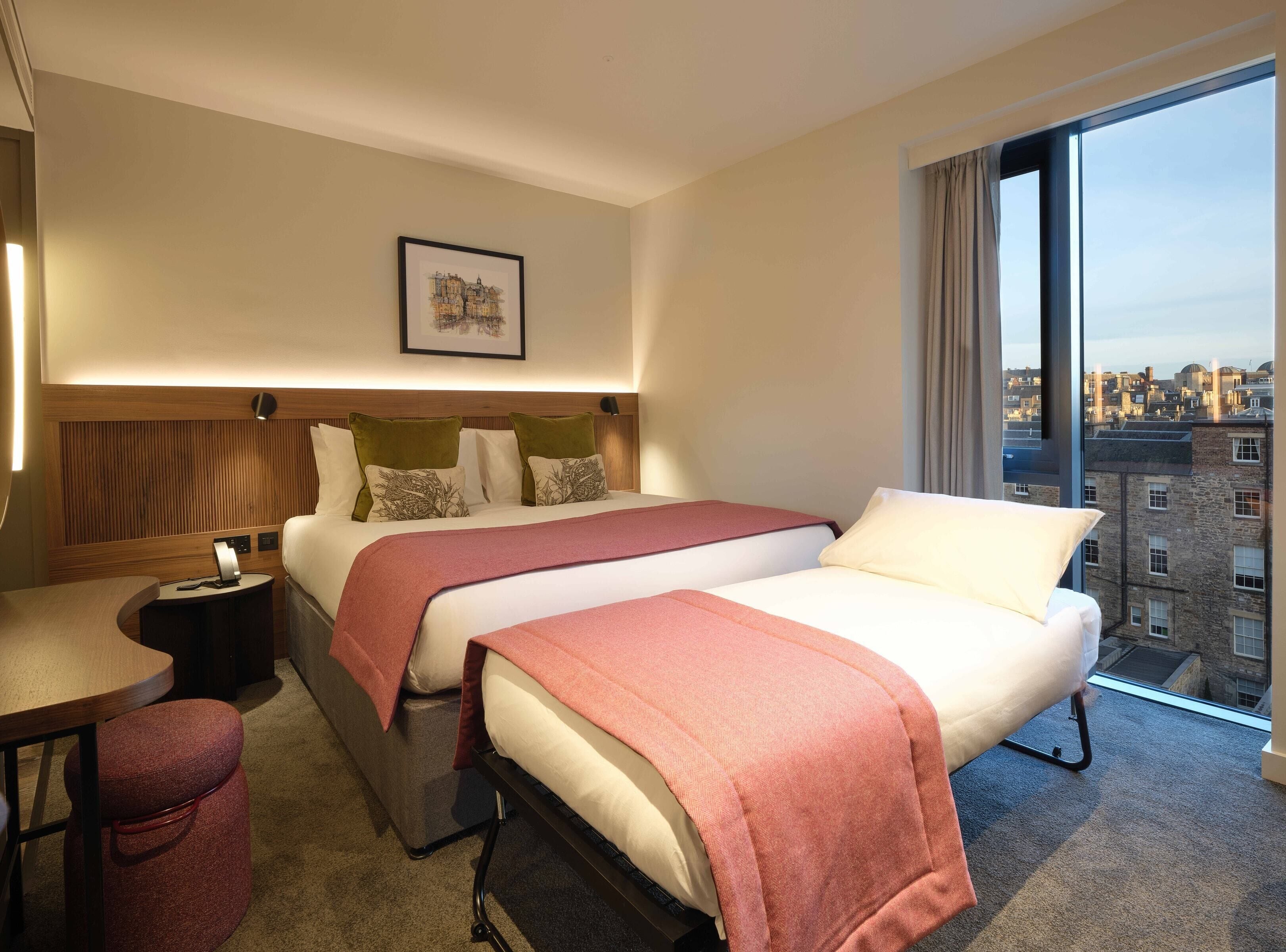 A hotel room with two beds, a desk, a chair, a nightstand, a framed picture, and a large window with a city view.