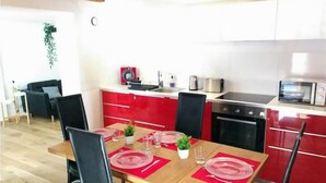 A dining area with a wooden table set for four, surrounded by black chairs. A red kitchen with white cabinets is visible in the background.