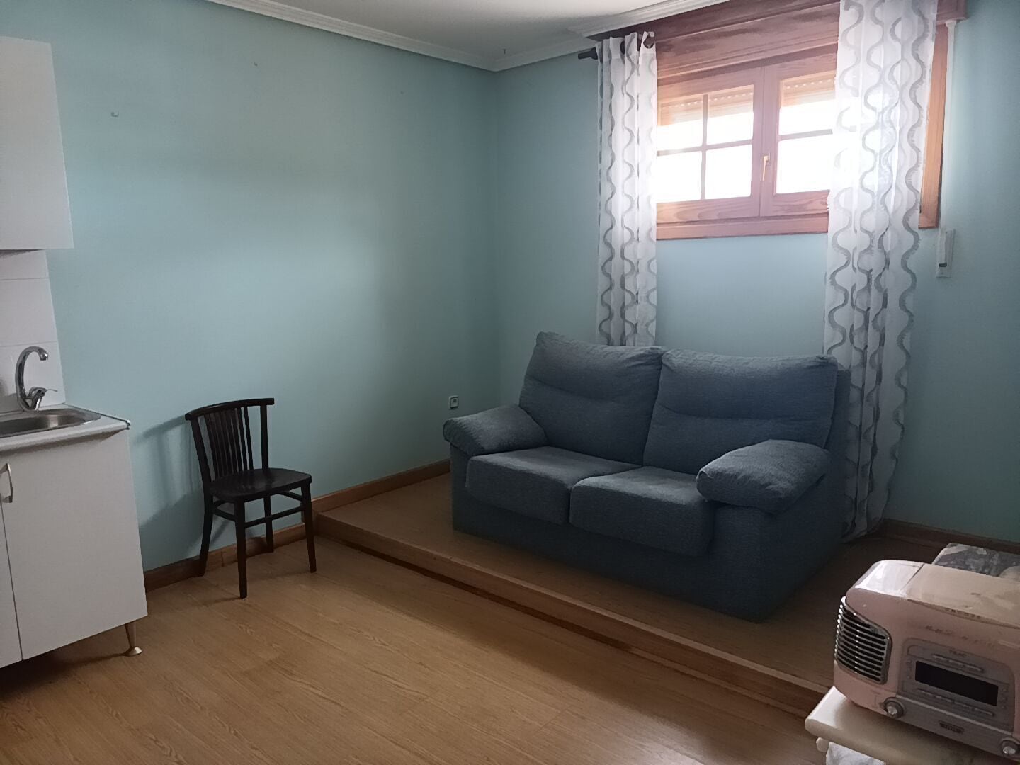 Una pequeña sala de estar con sofá, sillón, kitchenette y ventana con cortinas.