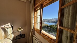 A room with a view of a lake and mountains, a radiator, a lamp, and a bed with floral bedding.