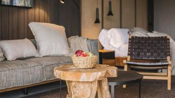 Una habitación acogedora con un sofá gris, una pequeña mesa redonda con una cesta de mimbre llena de fruta, una silla de madera y una cama con cabecera oscura.