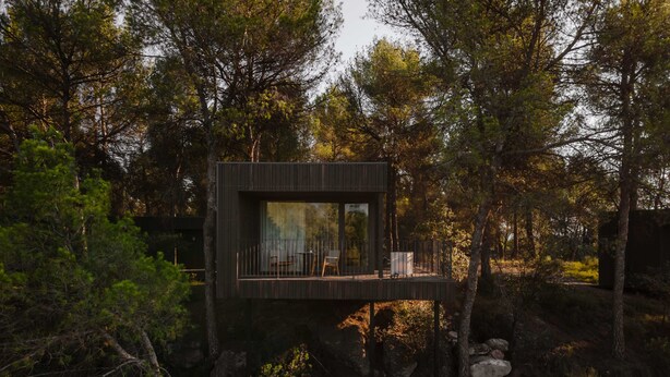 Een moderne houten chalet met een terras, omringd door bomen en rotsen.