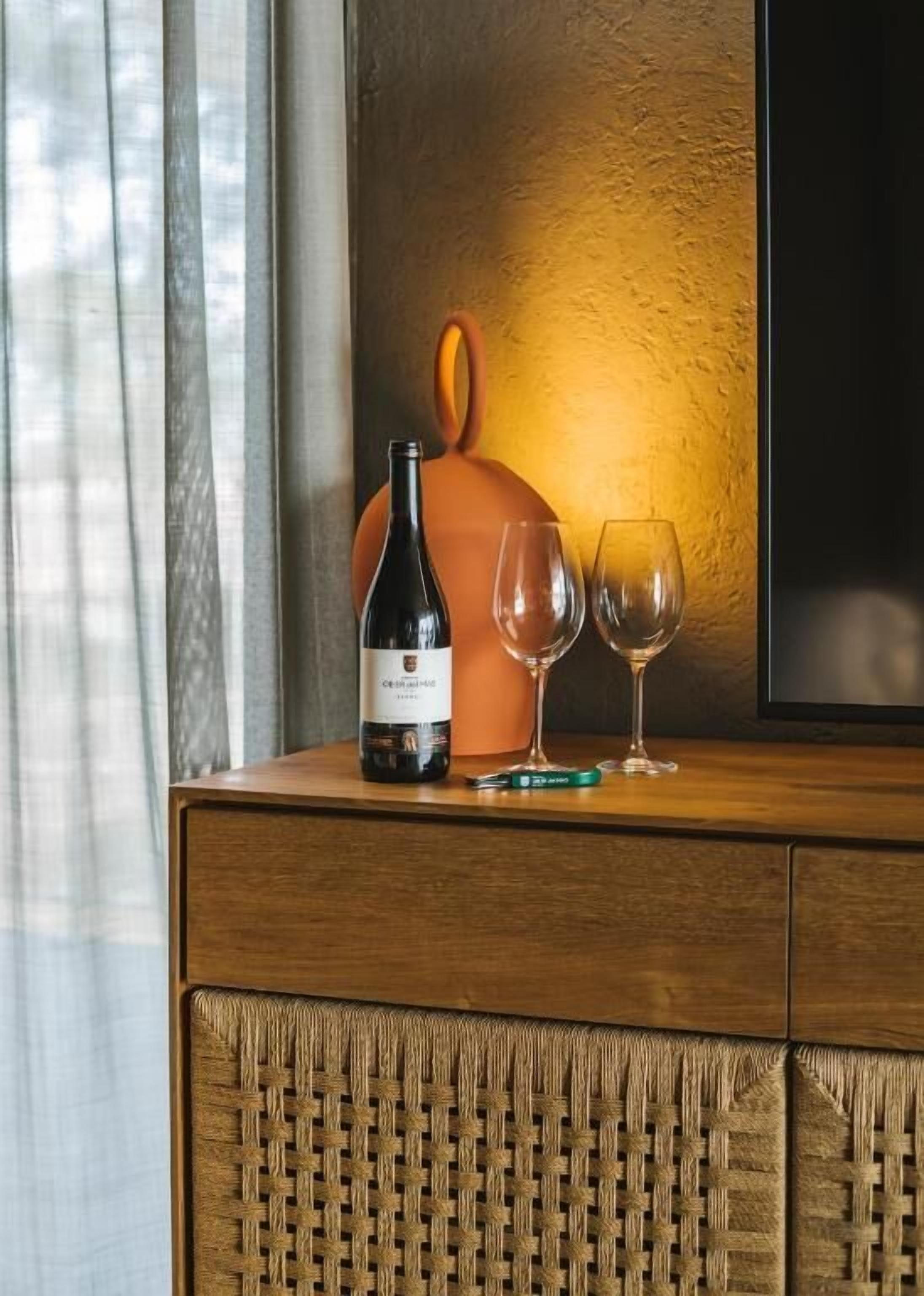 Een houten kastje met een gevlochten voorpaneel, een fles wijn en twee wijnglazen erbovenop.