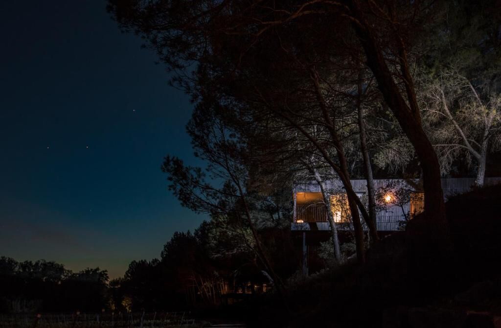 Una casa en árbol con luz en su interior, rodeada de árboles y una valla.