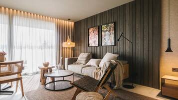 Un salon moderne avec un canapé, une table basse et des fauteuils. On y trouve une grande fenêtre avec des rideaux, un luminaire fixé au mur et deux tableaux encadrés.