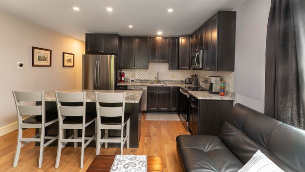 A compact kitchen with dark cabinets, a refrigerator, microwave, and a sink. There is a dining area with a table and chairs, and a black leather sofa. - Perfect Location in North Conway Village

 (Conway)