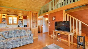 A cozy cabin interior with a wooden staircase, a sofa, a dining table, and a television. - Warm & Cozy Clayton Cabin Near St Lawrence River! (Clayton)