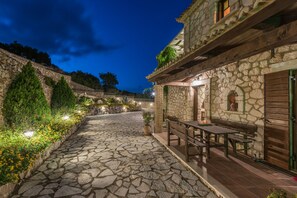 Un patio empedrado con un banco y una mesa de madera, rodeado de muros de piedra y vegetación.