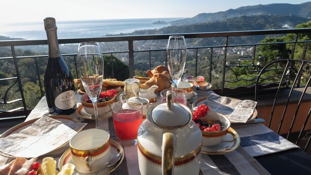 A table set with a bottle, glasses, and food items on a balcony overlooking a scenic view.
