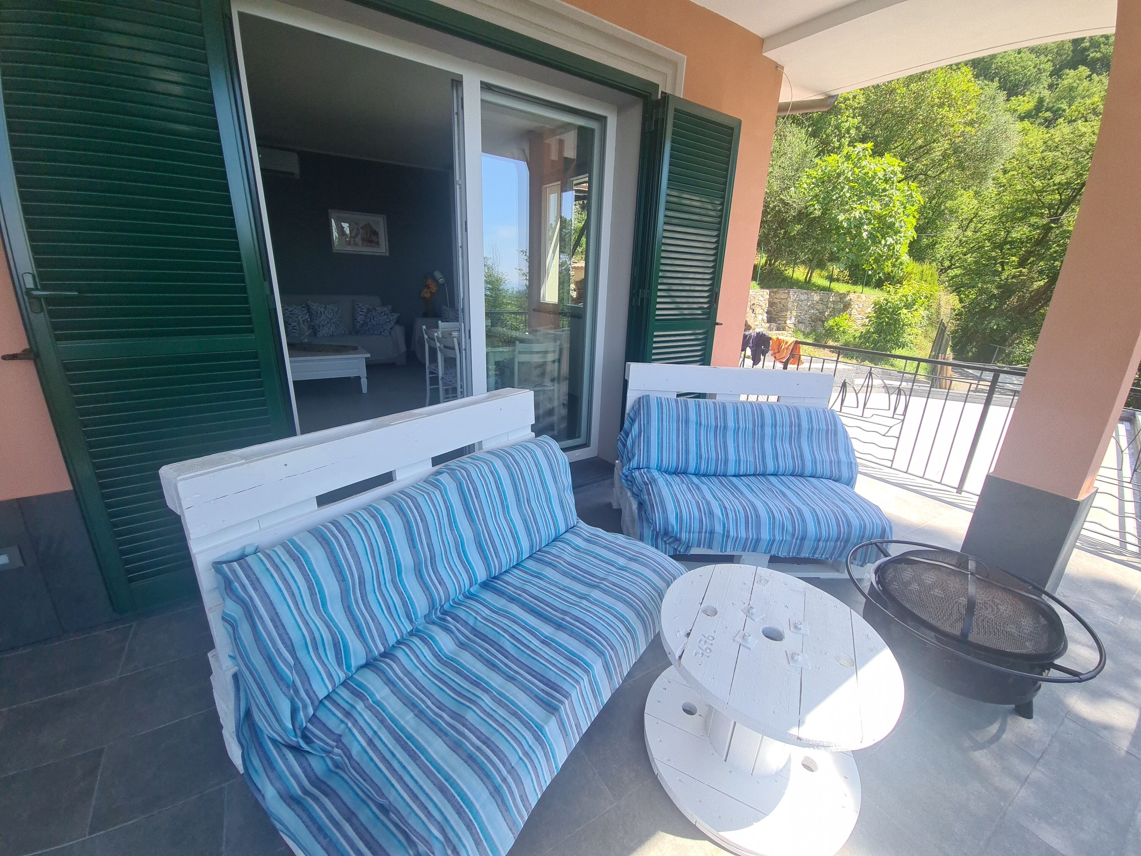 A balcony with two striped sofas and a round table, a sliding glass door, and a view of a garden.