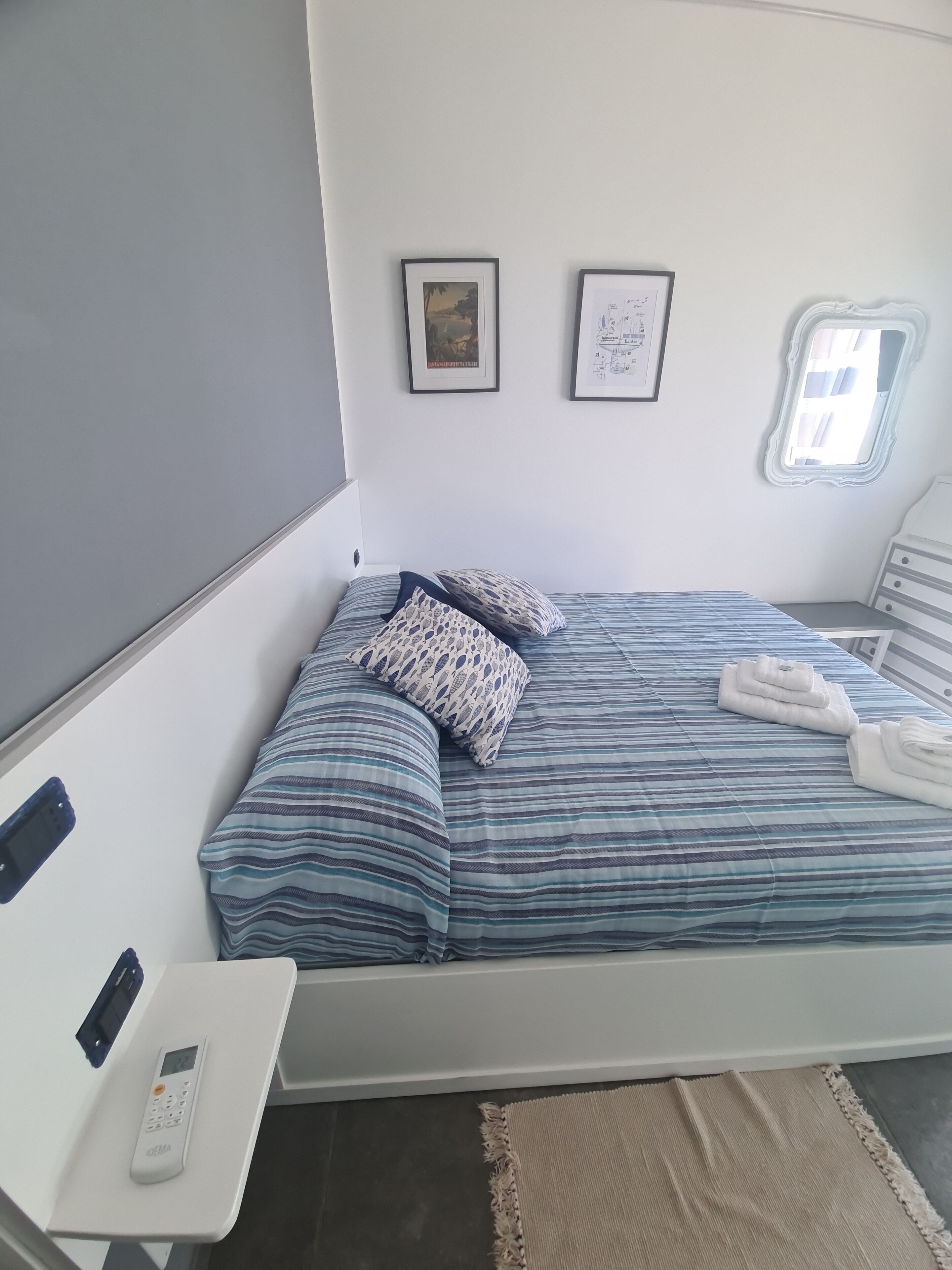 A neatly arranged bedroom with a bed, pillows, a bedside table with a phone and remote, a small window, and two framed pictures on the wall.