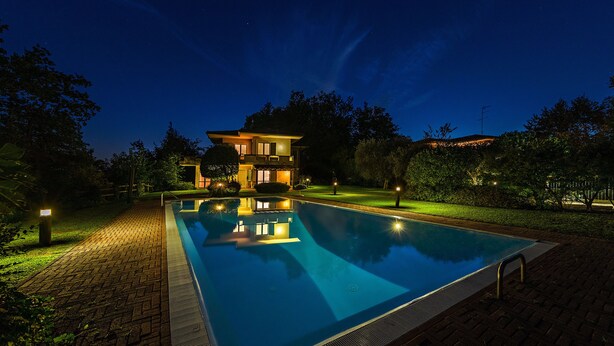 Una piscina con borde de ladrillo, rodeada por un césped iluminado y árboles.