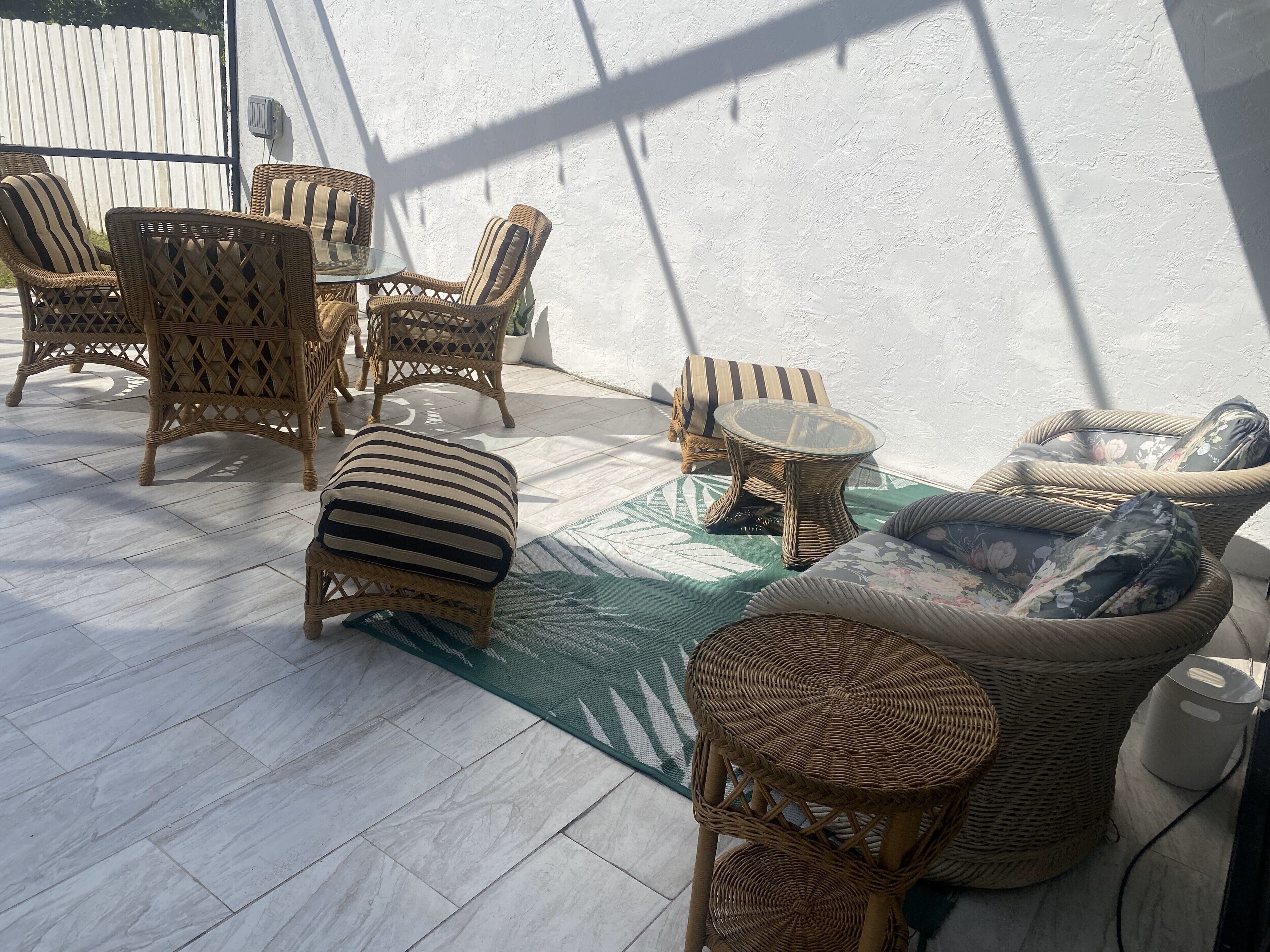 Un patio soleggiato con mobili in vimini, tra cui sedie, un tavolo e un divano, con un muro bianco e una recinzione in legno sullo sfondo.