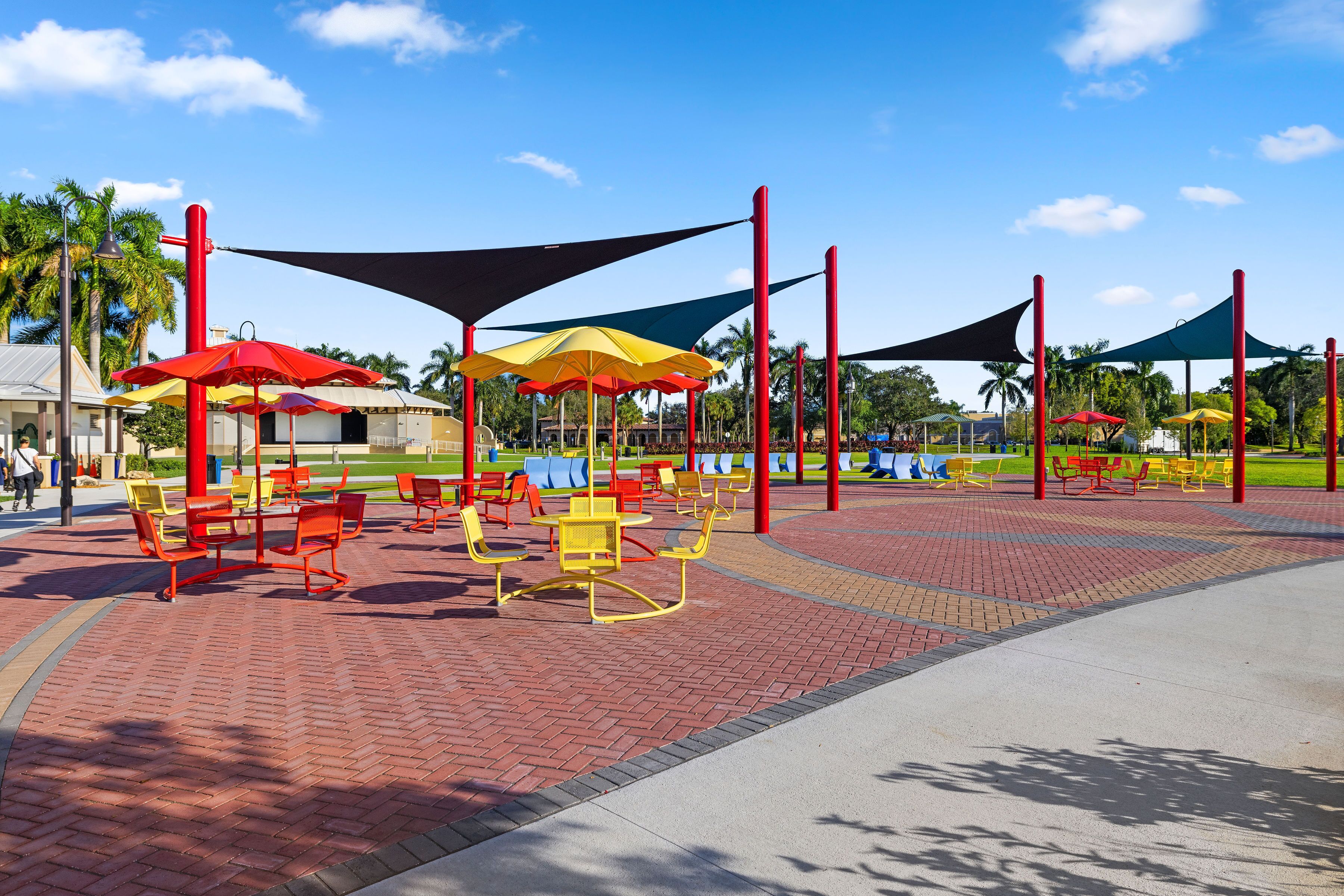 Un parque con zonas de estar al aire libre, que incluyen mesas y sillas, protegidas por sombrillas grandes sostenidas por postes rojos.