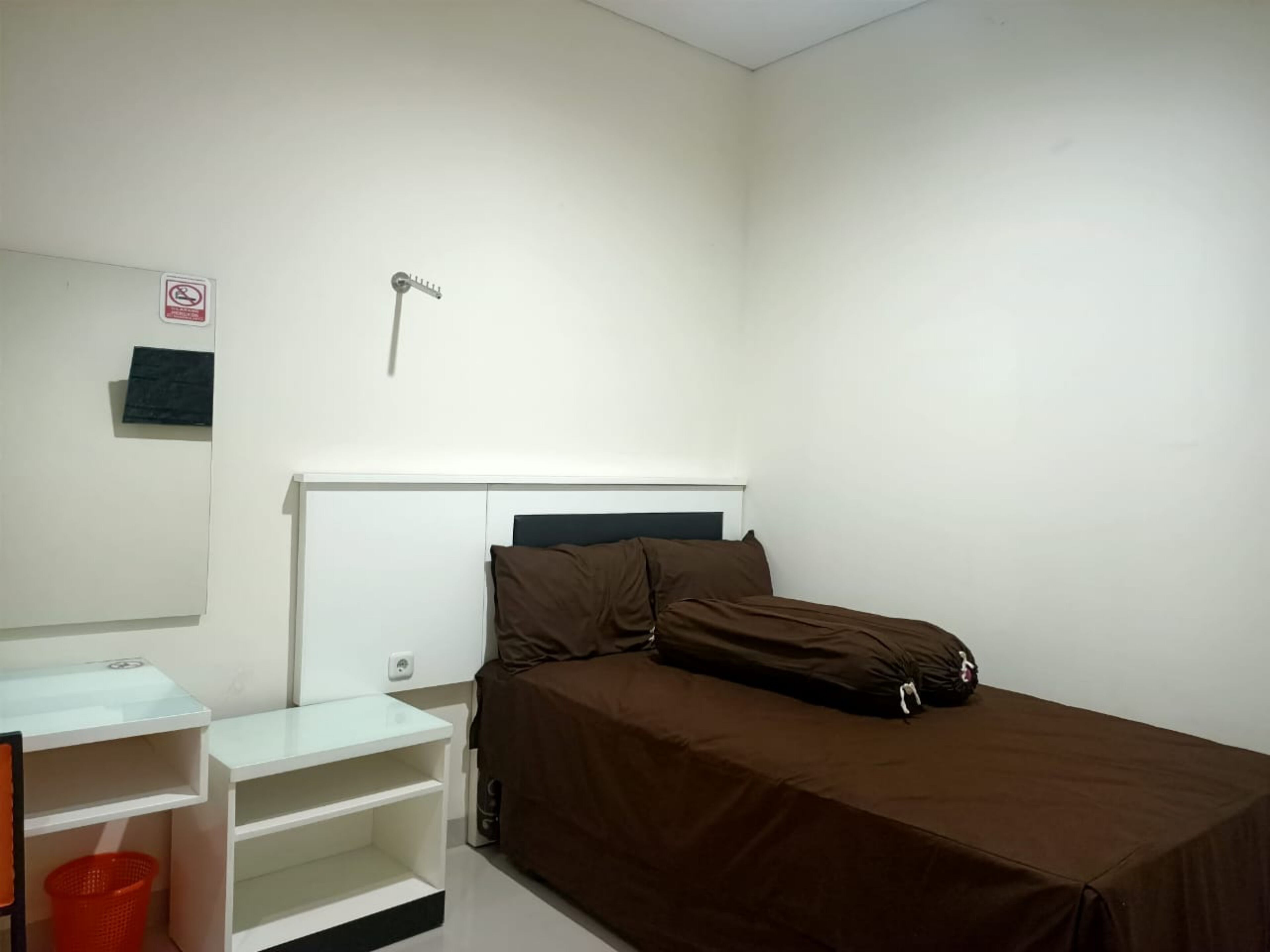 Una habitación pequeña y limpia con una cama, un escritorio y una silla. Hay una cesta roja en el suelo y una pared blanca con un cartel de prohibido fumar.