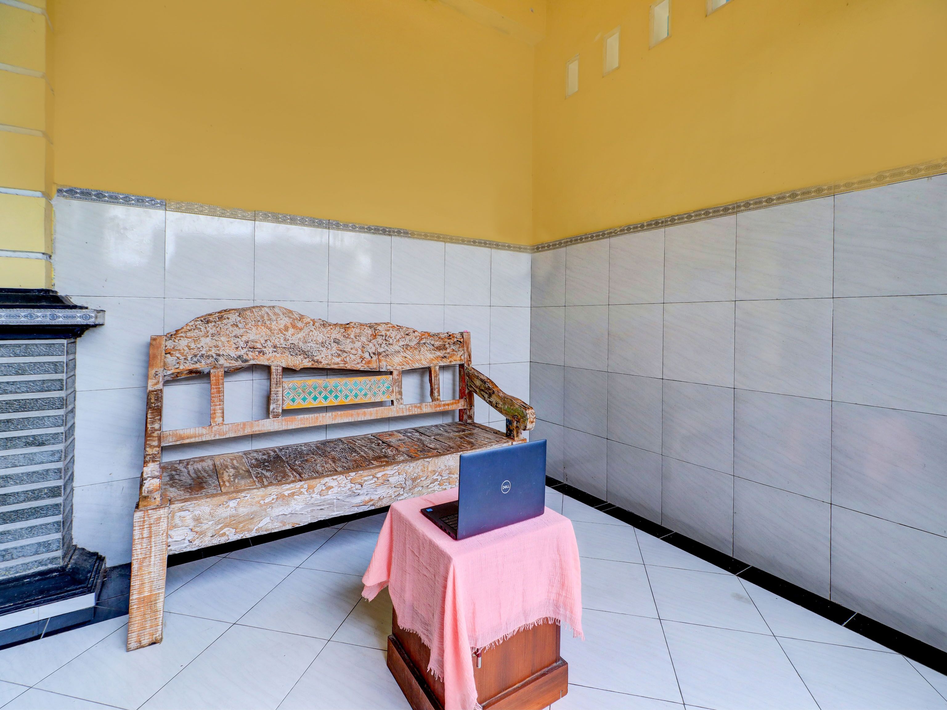 A room with a tiled floor, a bench with a laptop on a table, and a yellow wall.