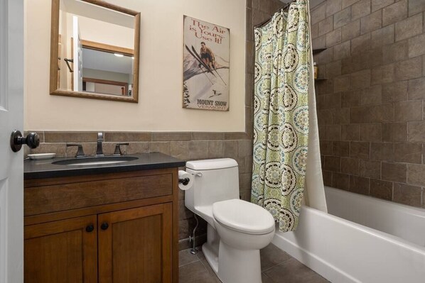 Une salle de bain avec un meuble-lavabo en bois, des toilettes, un lavabo, un miroir, un rideau de douche et une affiche accrochée au mur.