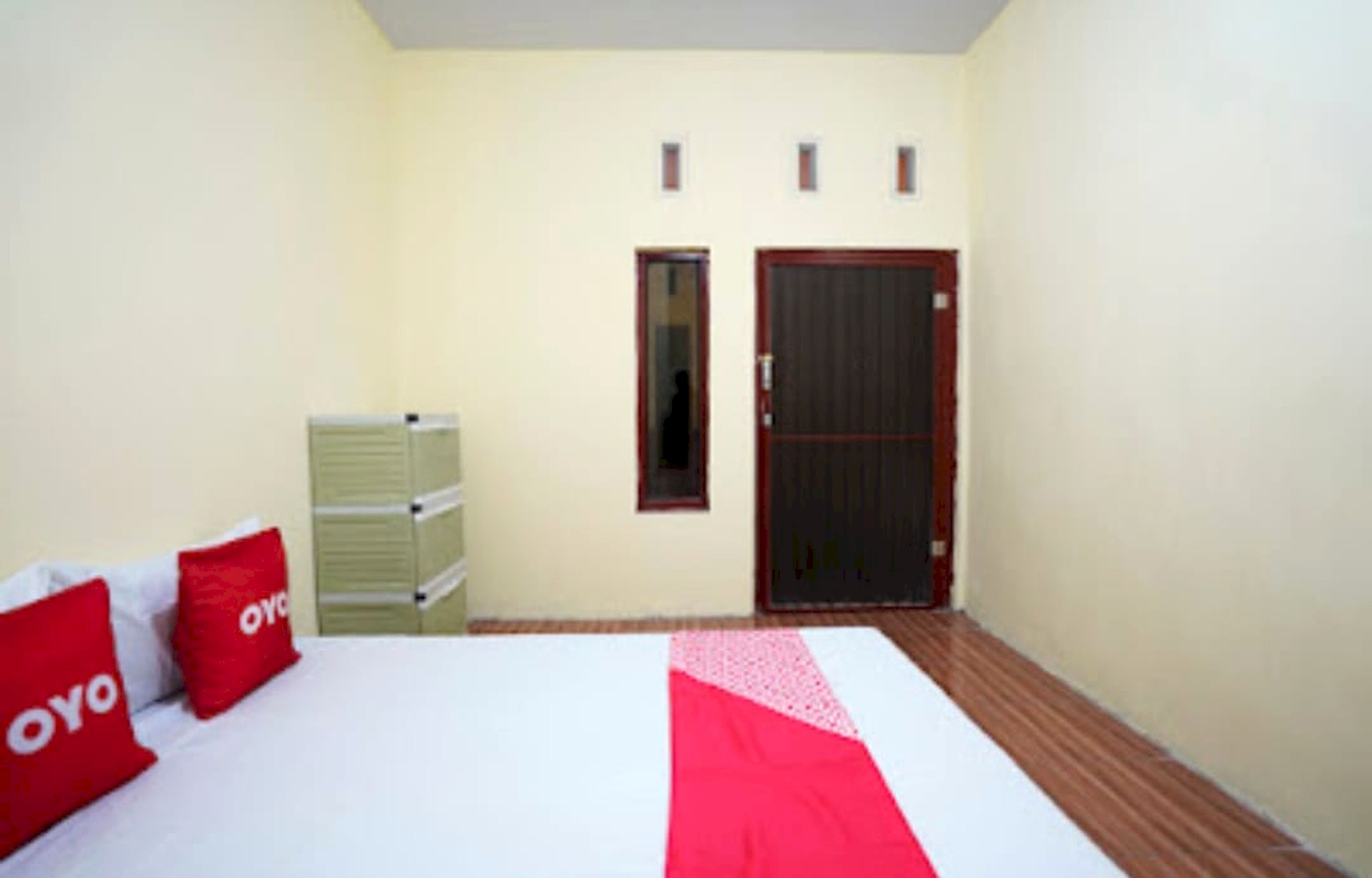 A bedroom with a bed, two red pillows, a wooden floor, a white and red bedspread, a green chest of drawers, and a dark door.