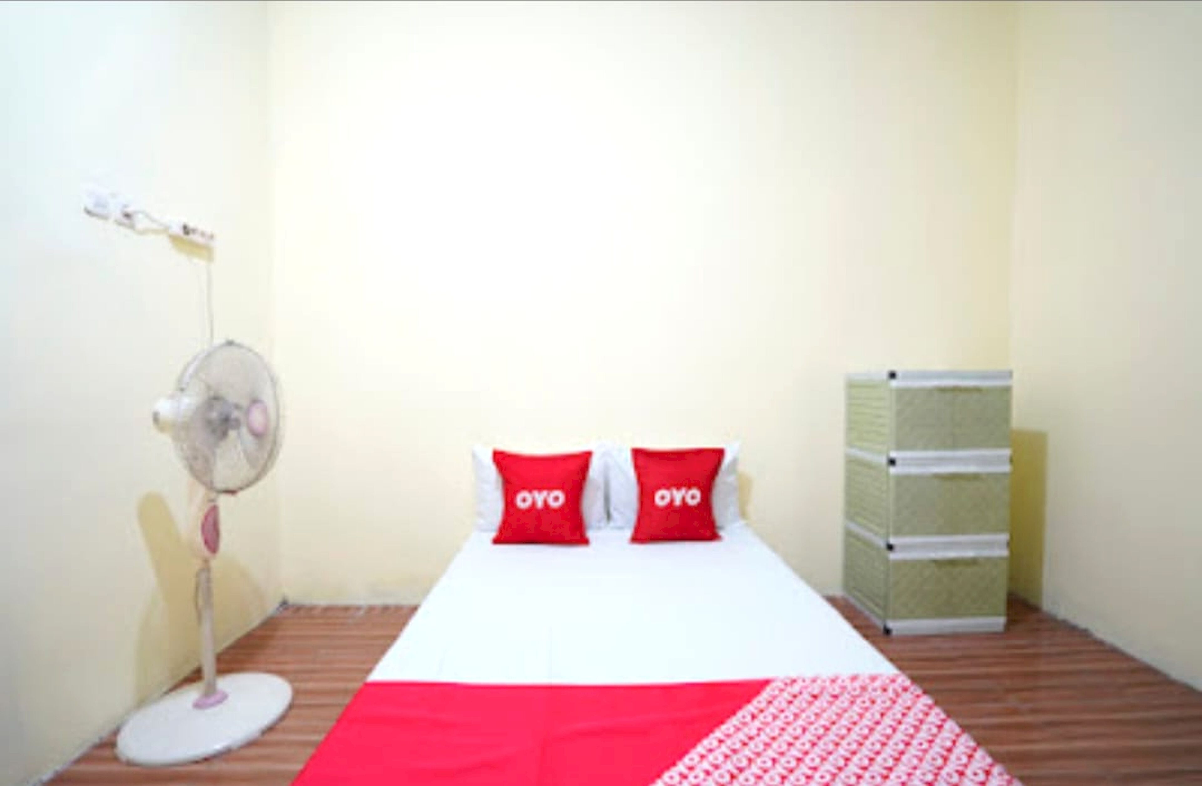 A small, simple room with a bed, two red pillows, a standing fan, and a stack of storage boxes.