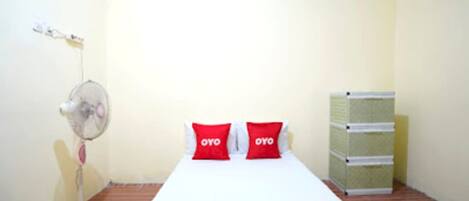 A small, simple room with a bed, two red pillows, a standing fan, and a stack of storage boxes.