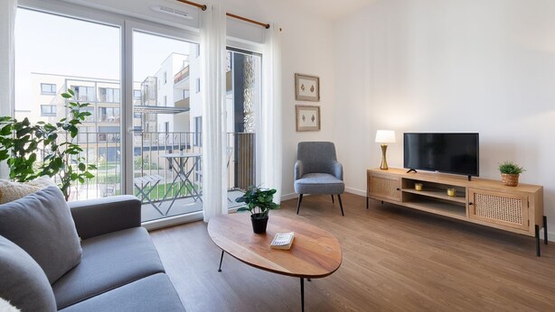 Un salon moderne avec un canapé, une table basse, un téléviseur et un balcon offrant une vue sur les immeubles.