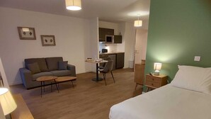 Une chambre d’hôtel comprenant un lit, un canapé, une table à manger avec des chaises, un petit coin cuisine et un bureau avec une lampe.