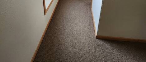 Un pasillo estrecho con pisos alfombrados, detalles de madera y una cama con colcha estampada a lo lejos.