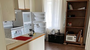 A compact kitchen with a stove, microwave, and refrigerator. There's a wooden cabinet and a small bookshelf with books and a plant.