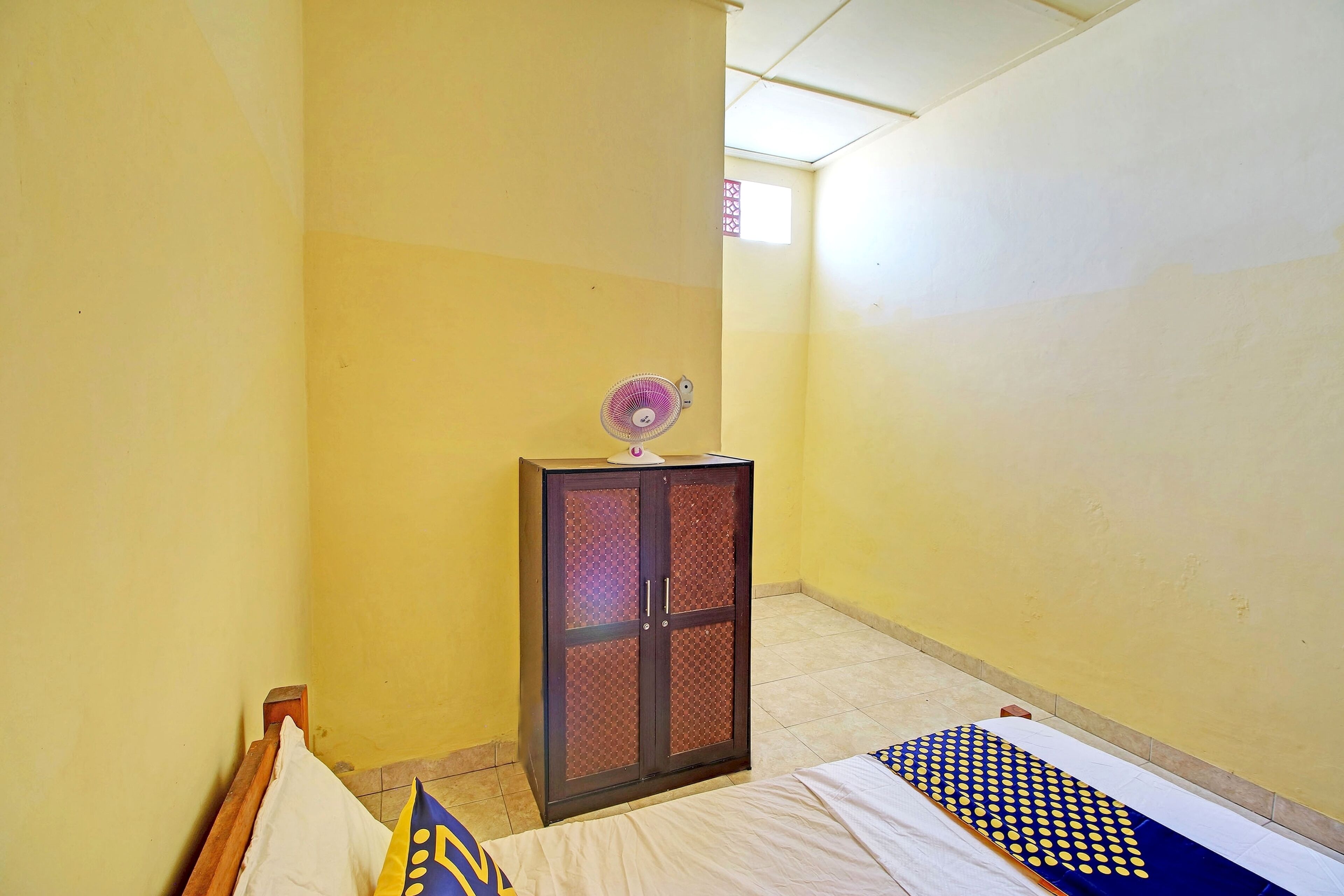 A small, narrow room with yellow walls, a bed with a patterned bedspread, a wooden headboard, and a cabinet with a fan on top.