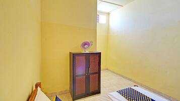 A small, narrow room with yellow walls, a bed with a patterned bedspread, a wooden headboard, and a cabinet with a fan on top.