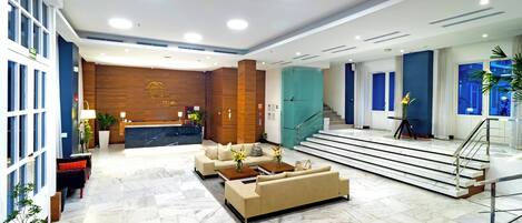 A modern hotel lobby with a reception desk, sofas, a coffee table, and a staircase leading to an upper floor.