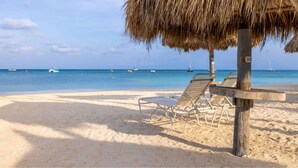 Une plage avec une structure surmontée d’un toit de chaume, un poteau en bois portant une enseigne et plusieurs chaises longues vides.