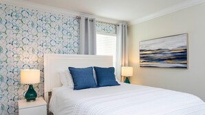 Ein ordentlich bezogenes Bett mit weißer Bettwäsche und blauen Kissen, ein weißer Nachttisch mit einer grünen Lampe und eine Wand mit blau-weißem abstrakten Muster.