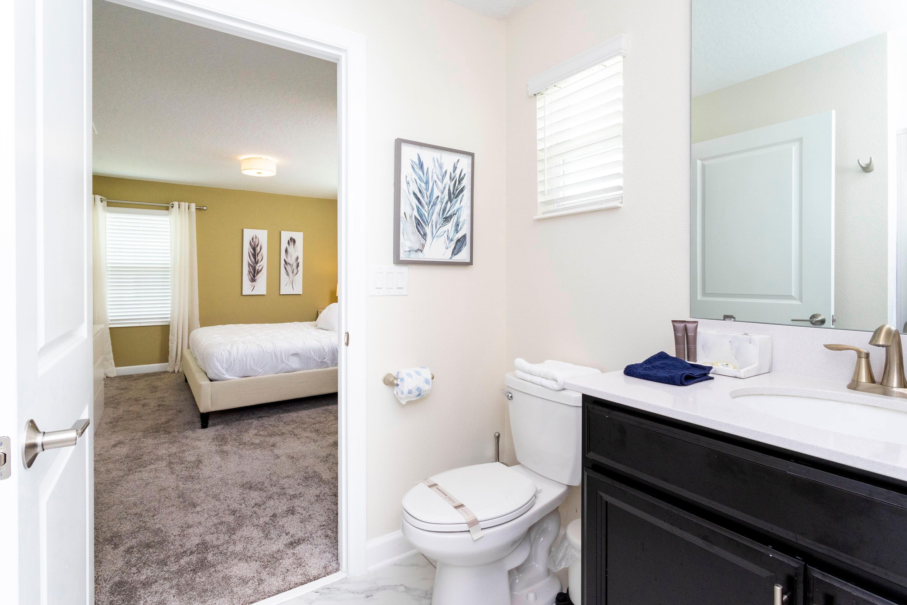 A bathroom with a toilet, sink, and mirror. There is a bed in the adjacent room.