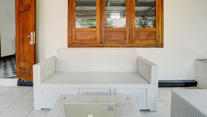 A white woven sofa with a laptop on it, a wooden door, and a window with a view of greenery. - SPOT ON 93014 Griya Nautika Sukodono (Sidoarjo)
