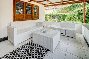 Ein überdachter Außenbereich mit weißer Rattan-Sitzgruppe, einem Ottomanen, einem gemusterten Teppich und Blick auf Grünflächen.