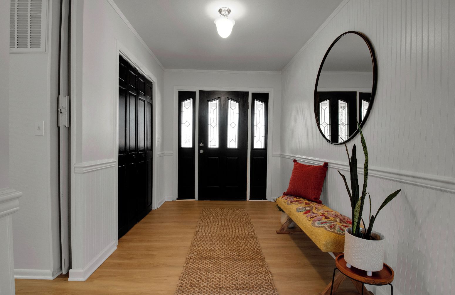 A hallway with a wooden floor, a bench with a patterned cushion, a potted plant, and a mirror on the wall.