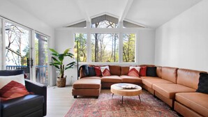 A spacious living room with a large L-shaped sectional sofa, a round coffee table, and a black armchair. There is a large window with a view of trees, and a potted plant is visible.