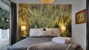 A bedroom with a large bed, two bedside lamps, a forest-themed wallpaper, and a view of a balcony.