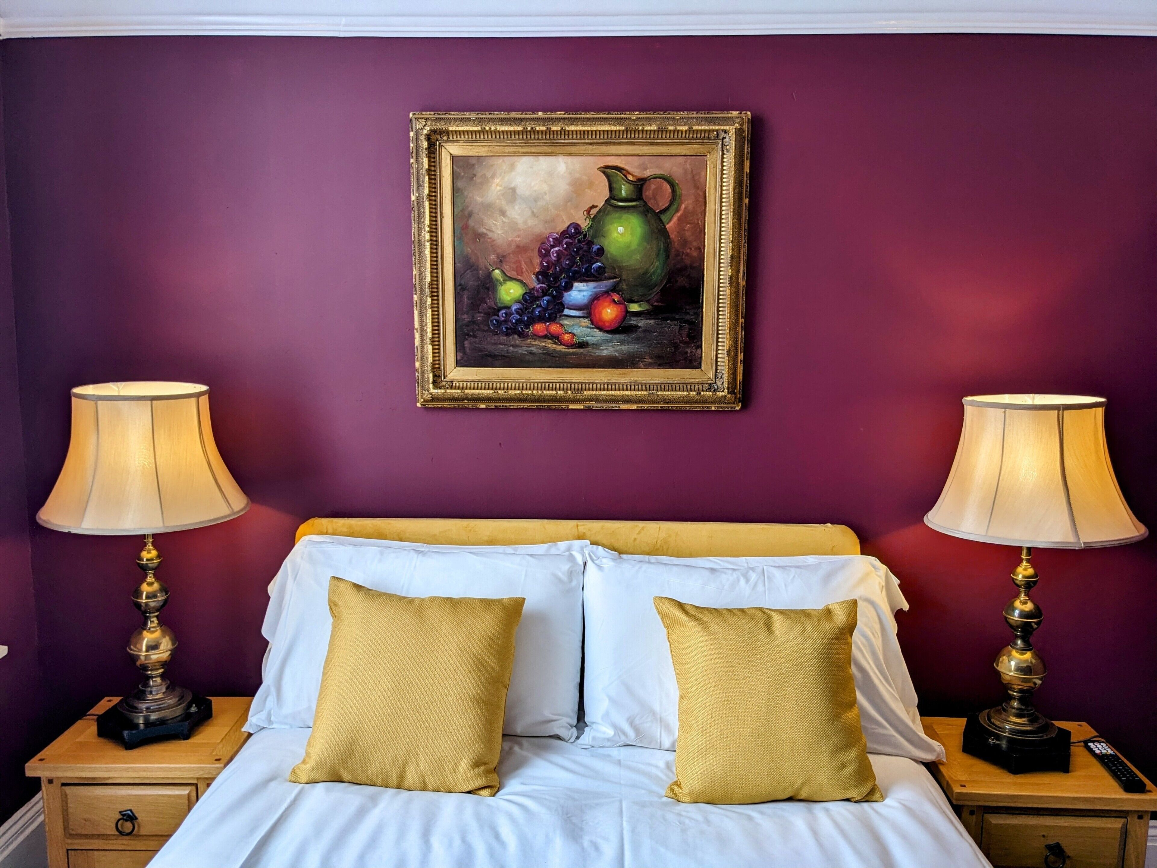 A bedroom with a bed, two nightstands, two lamps, a painting of fruit, and a purple wall.
