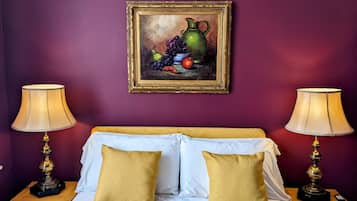 A bedroom with a bed, two nightstands, two lamps, a painting of fruit, and a purple wall.