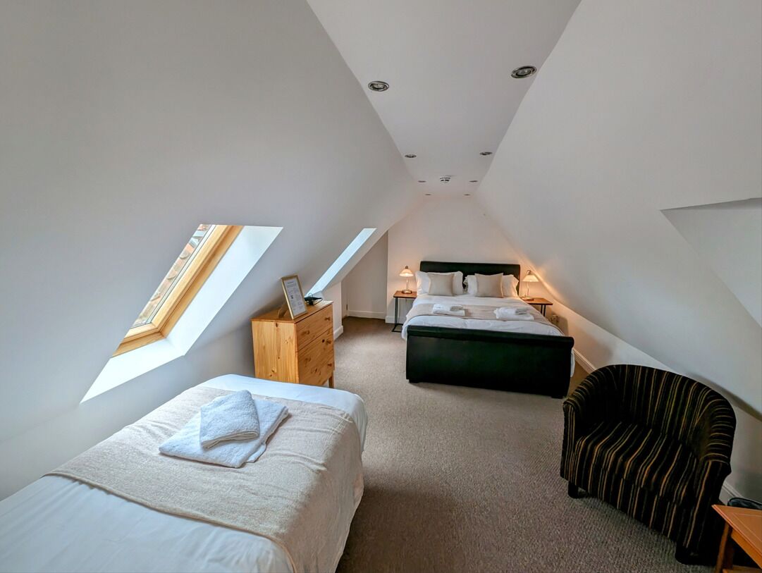 A modern, well-lit attic bedroom with a skylight, two beds, a chair, and a bedside table.